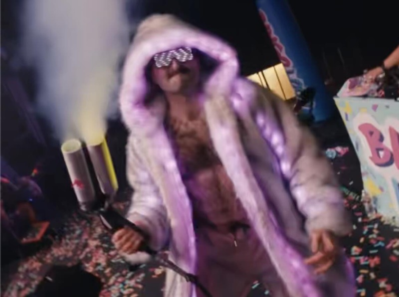 Scott Rowan as Max Chaos in a light-up fur coat and LED glasses, surrounded by confetti and smoke